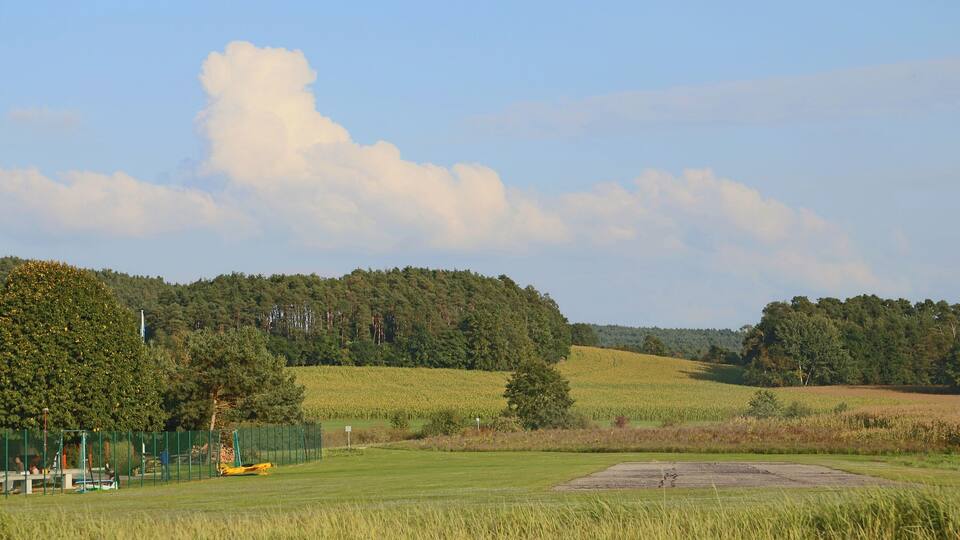 A grassy field with a yellow golf cart, a fenced area, and a hill with trees in the background.