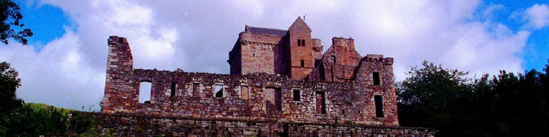 Un castillo de piedra histórico con ruinas, rodeado de abundante césped verde y árboles.