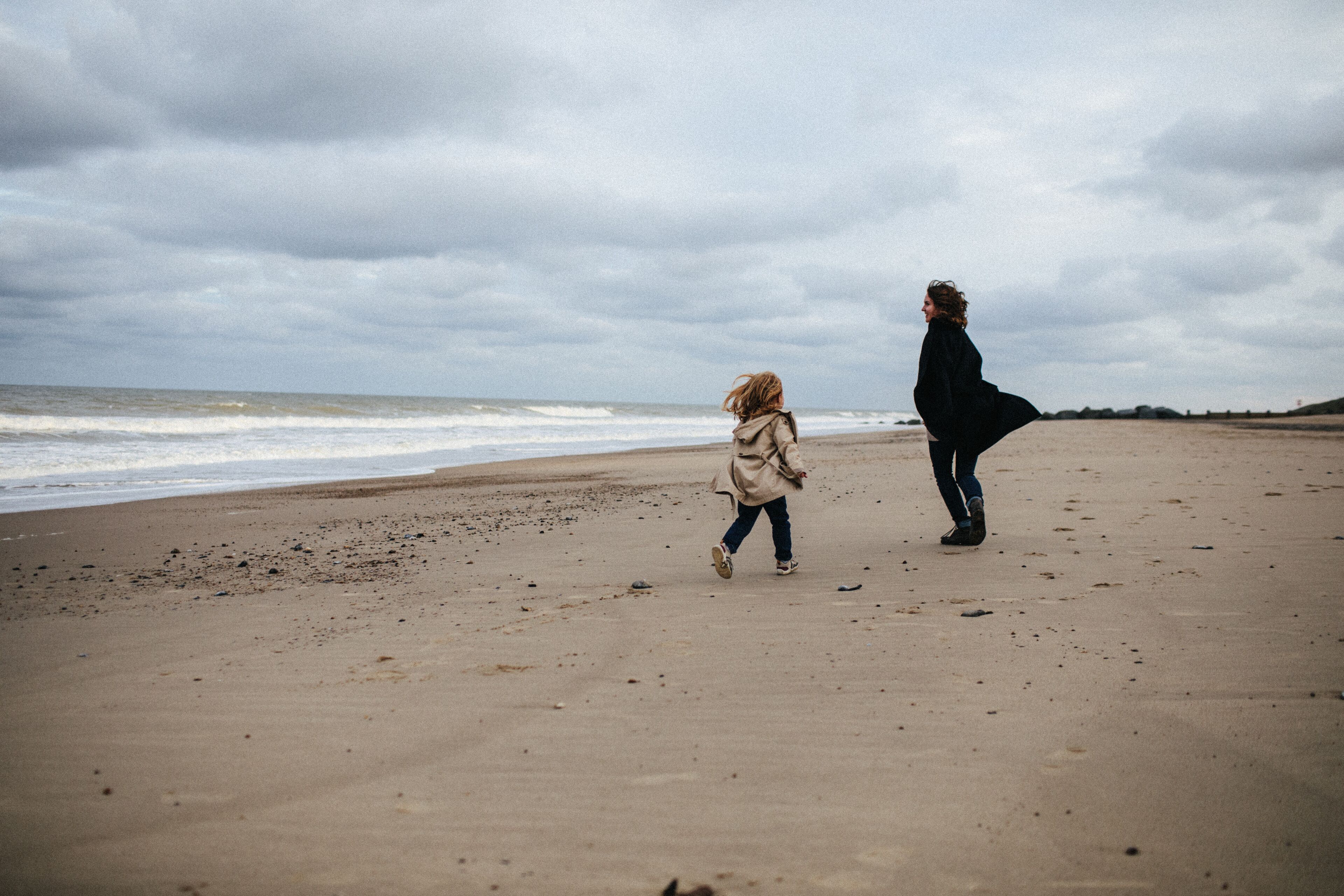 Mother in a warm coat and little daughter in a trench coat run after each other and have fun on the sandy beach with a strong wind in autumn, Norfolk, England