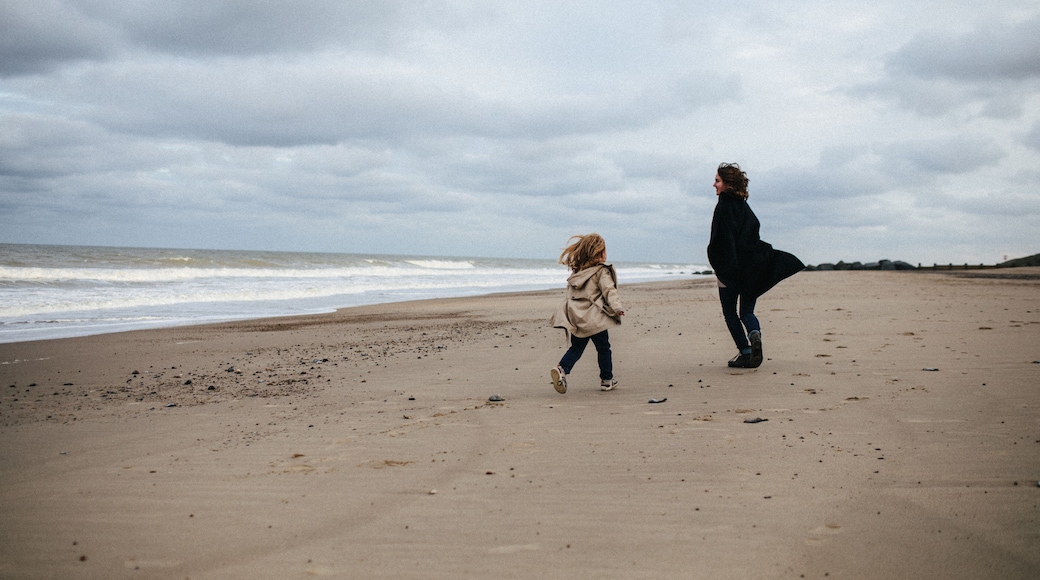 Mother in a warm coat and little daughter in a trench coat run after each other and have fun on the sandy beach with a strong wind in autumn, Norfolk, England