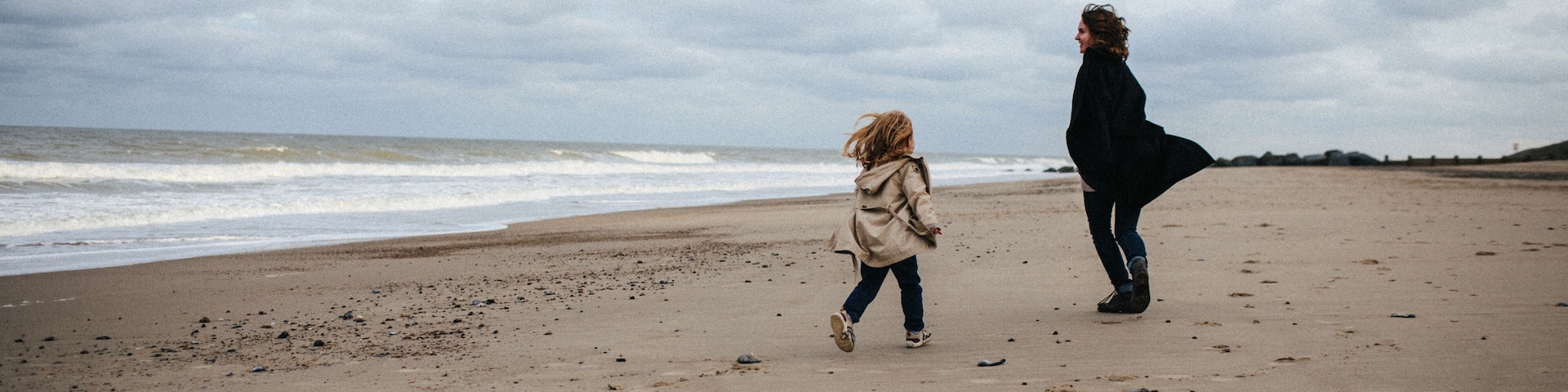 Mother in a warm coat and little daughter in a trench coat run after each other and have fun on the sandy beach with a strong wind in autumn, Norfolk, England