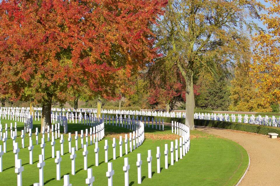 Un cimetière où des croix blanches sont disposées en rangées, entouré d’arbres aux feuilles d’automne.