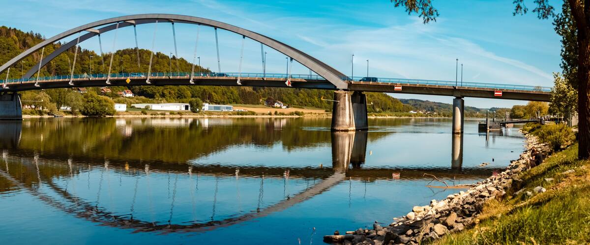 A bridge over a calm river with a reflection, surrounded by greenery and hills.