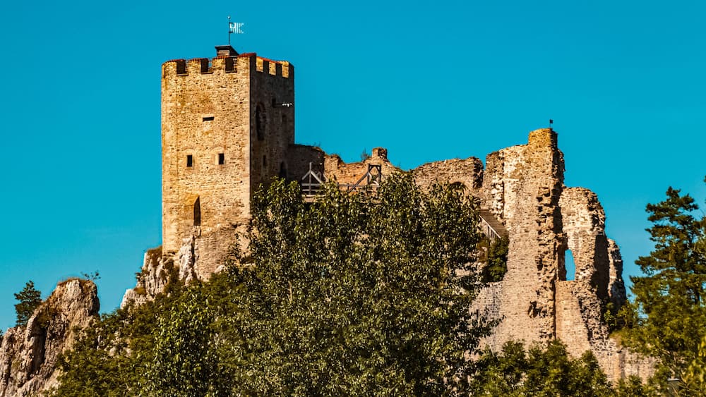Un château de pierre historique, doté d’une tour imposante, entouré d’une végétation luxuriante et d’un paysage rocheux.