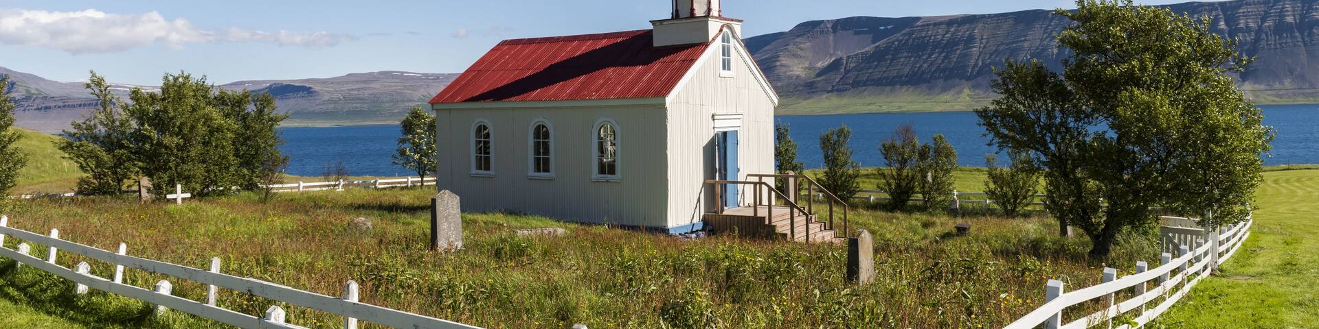 A white church with a red roof and a bell tower, surrounded by a white fence, near a lake with mountains in the background.
