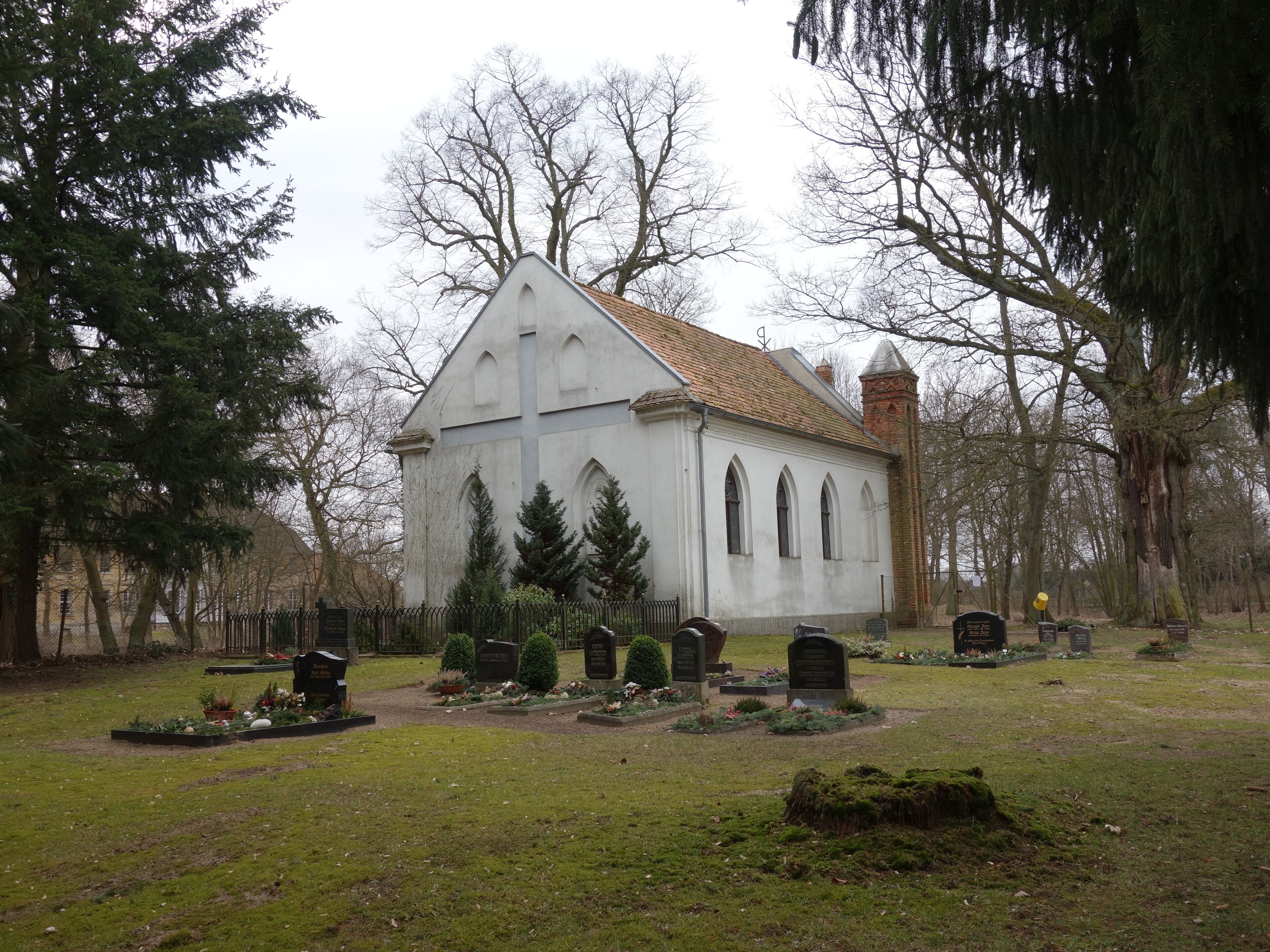 A white church with a tiled roof surrounded by trees and a cemetery.
