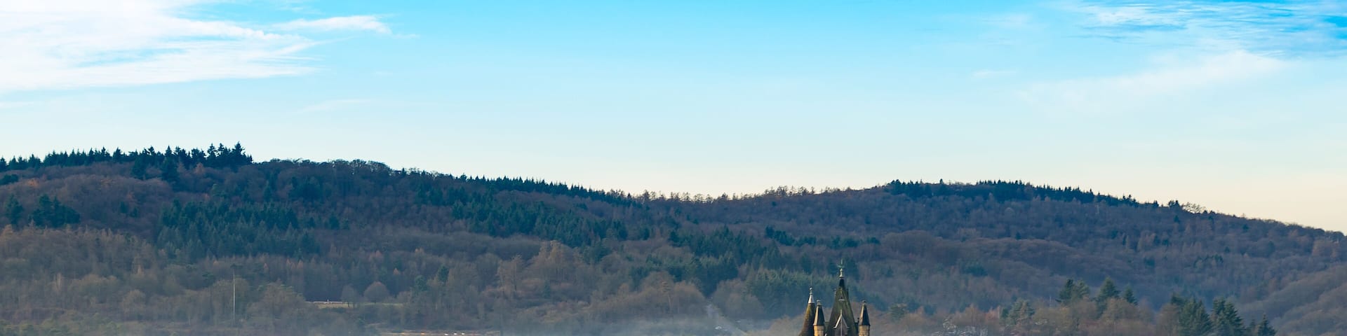 A historic castle with a tall tower, surrounded by a forested landscape and a misty valley.