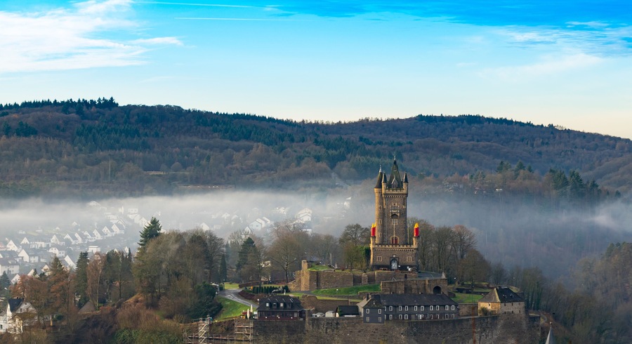 Eine historische Burg mit hohem Turm, umgeben von bewaldeter Landschaft und einem nebelverhangenen Tal.