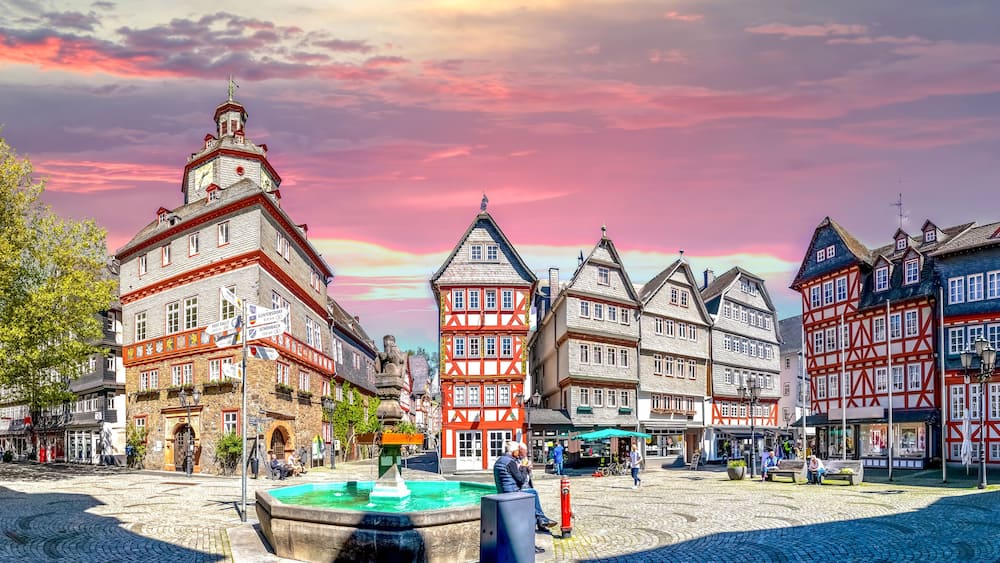 A historic European town square with cobblestone streets, traditional half-timbered buildings, and a fountain.