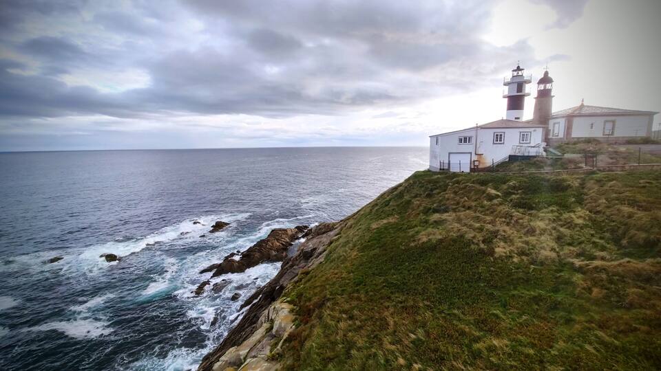 A coastal scene with a lighthouse and a white building on a grassy cliff overlooking the ocean.