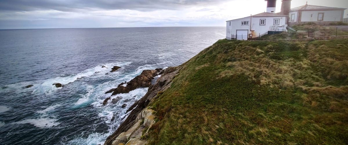A coastal scene with a lighthouse and a white building on a grassy cliff overlooking the ocean.