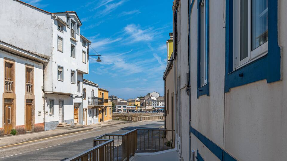 A narrow street with buildings on either side, a clear blue sky, and a distant view of a town.