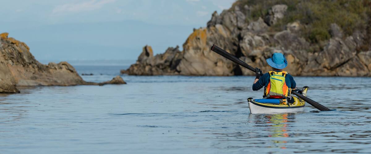 A kayaker in a small boat on calm water with a rocky coastline and a mountain in the background.