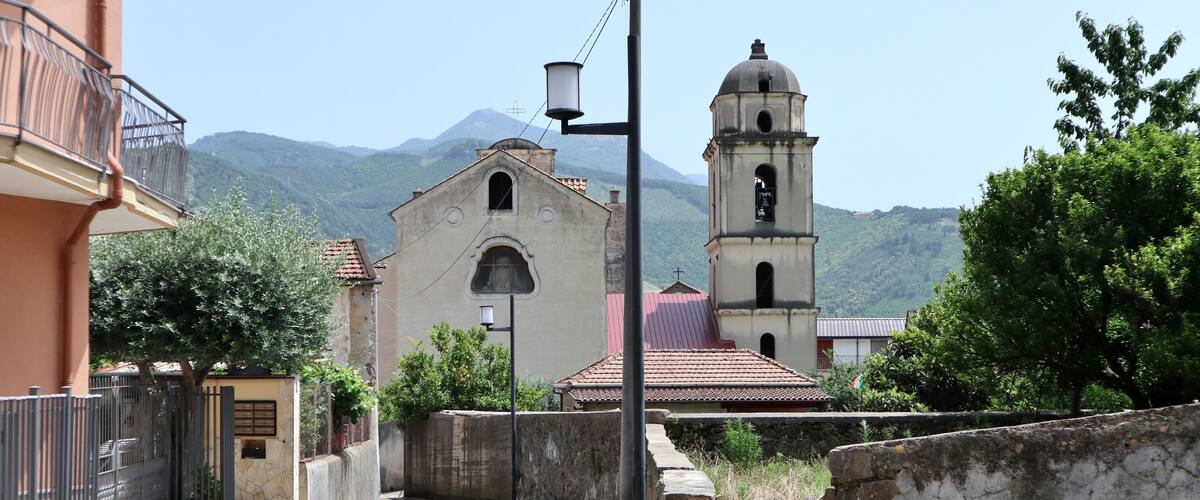 A narrow street leading to a church with a bell tower, surrounded by stone walls and greenery, with mountains in the background.