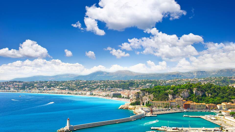 A coastal cityscape with a clear blue sky, a prominent lighthouse, and a marina with boats.