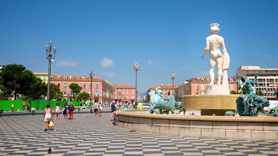 A public square with a fountain featuring a statue of a man and horses, surrounded by people and buildings.