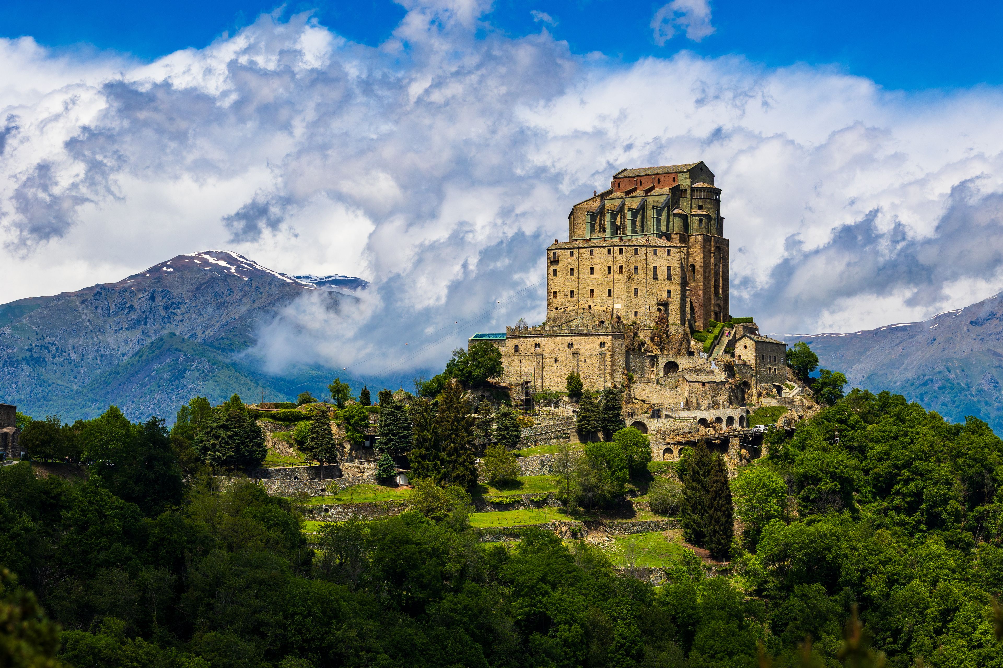 Sacra di San Michele abbey perched on Mount Pirchiriano at the entrance of the Susa Valley in spring, with the still snow-capped Alpine peaks emerging through the clouds in the background