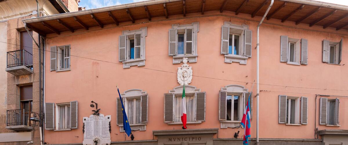A building with the sign MVNIFICIO, featuring multiple windows with shutters, a balcony, and flags.