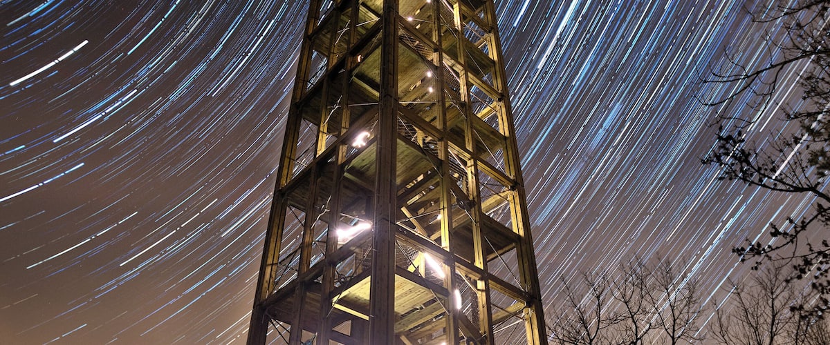A tall metal structure with lights at the top, surrounded by trees and a star trail in the night sky.