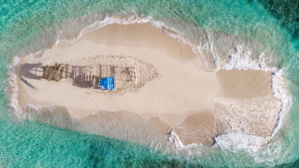 An aerial view of a beach with clear turquoise waters, a sandy shore, and a structure with a blue tarp.