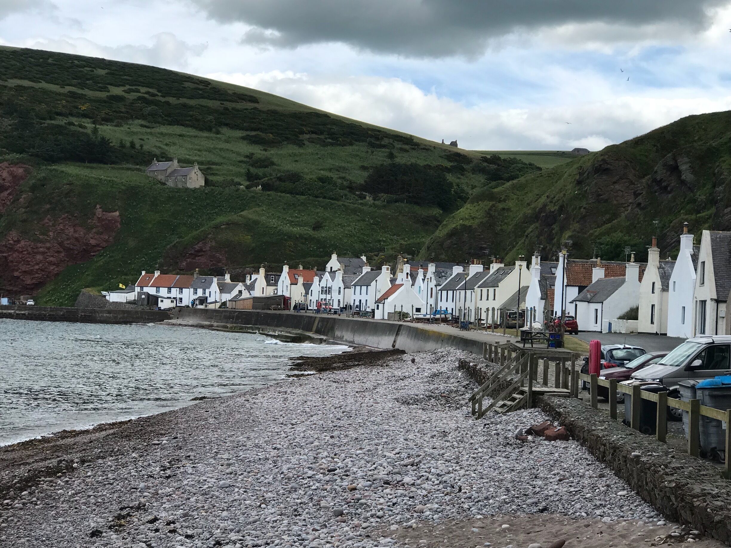 A coastal village with white houses along the shore, a rocky beach, and green hills in the background.