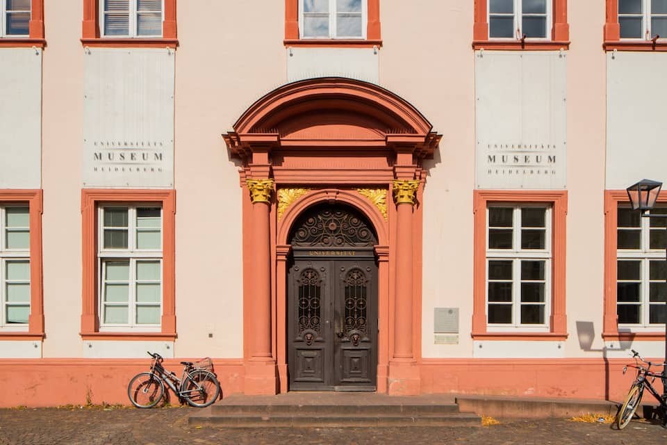 Ein pinkfarbenes Gebäude mit einer schwarzen Tür, zwei Fahrräder sind voran geparkt, und ein Schild mit der Aufschrift "Universitats Museum Heidelberg".