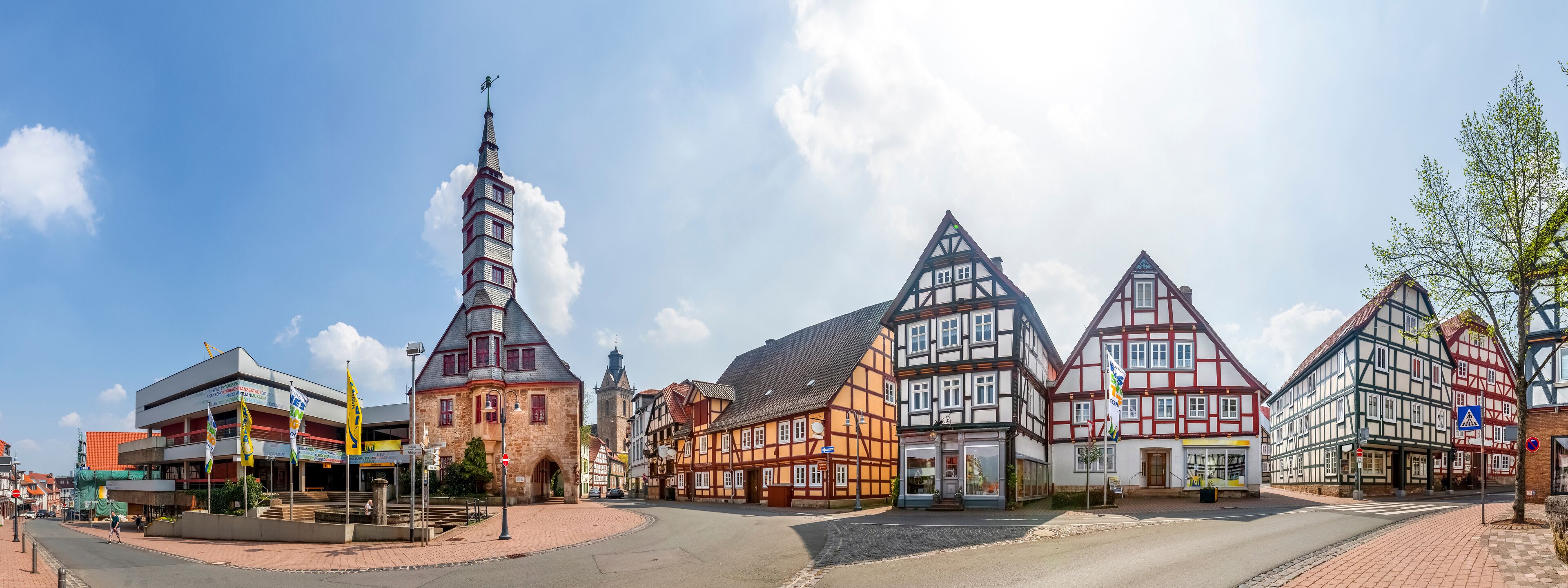 A historic town square with traditional half-timbered houses, a church tower, and a modern building with flags.