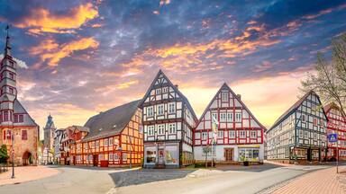 Uma cidade europeia com casas tradicionais de enxaimel, uma torre de igreja e um edifício moderno com bandeiras.