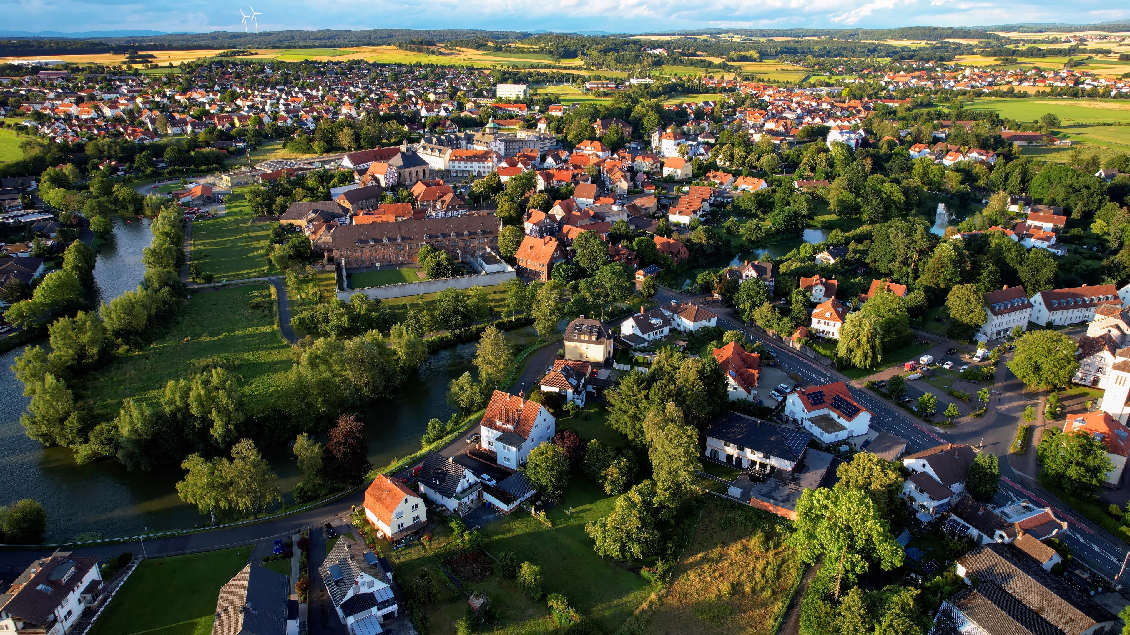 An aerial view of a town with a river, green fields, and wind turbines in the distance.