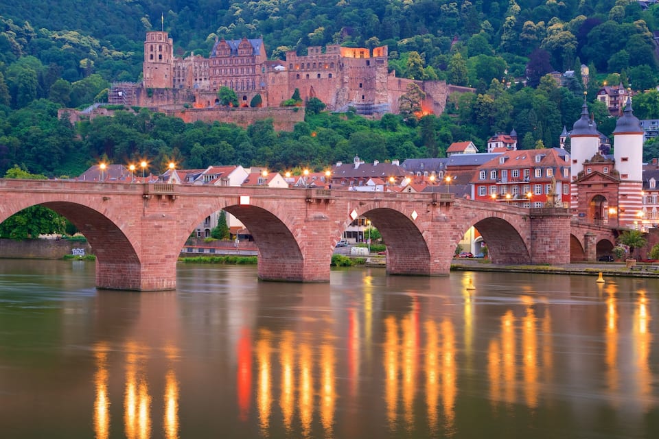 Eine Stebrücke über einem Fluss, im Hintergrund ein Schloss.