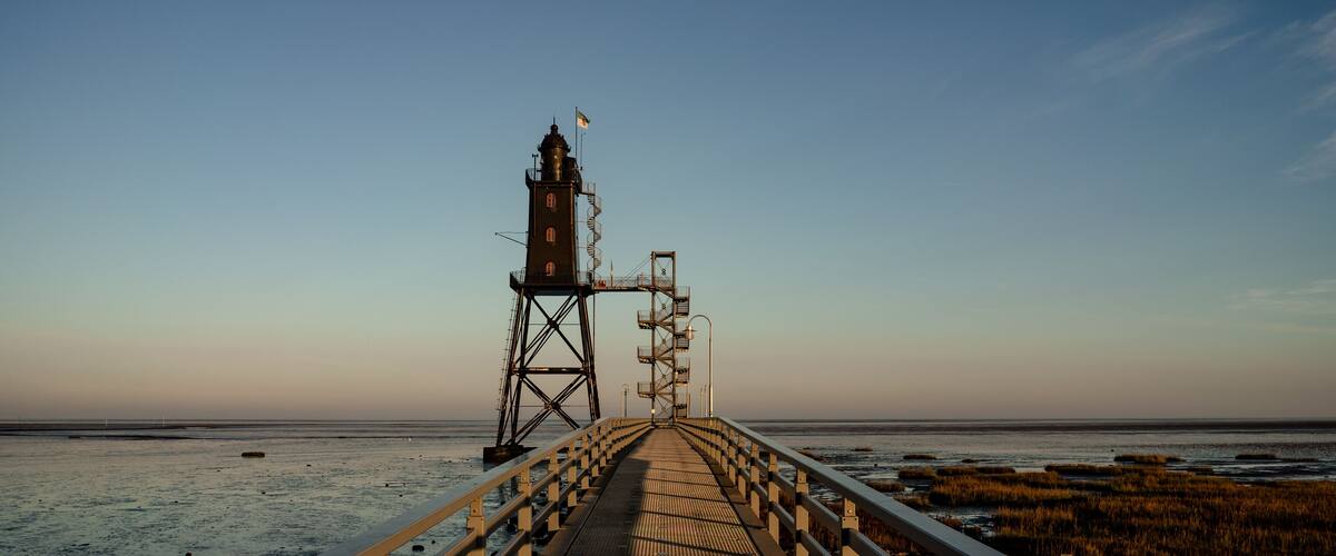 Een steiger die naar een vuurtoren leidt, met een vlag, een heldere lucht, en een strand met kweldergebied.