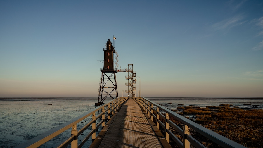 A pier leading to a lighthouse with a flag, a clear sky, and a beach with marshland.