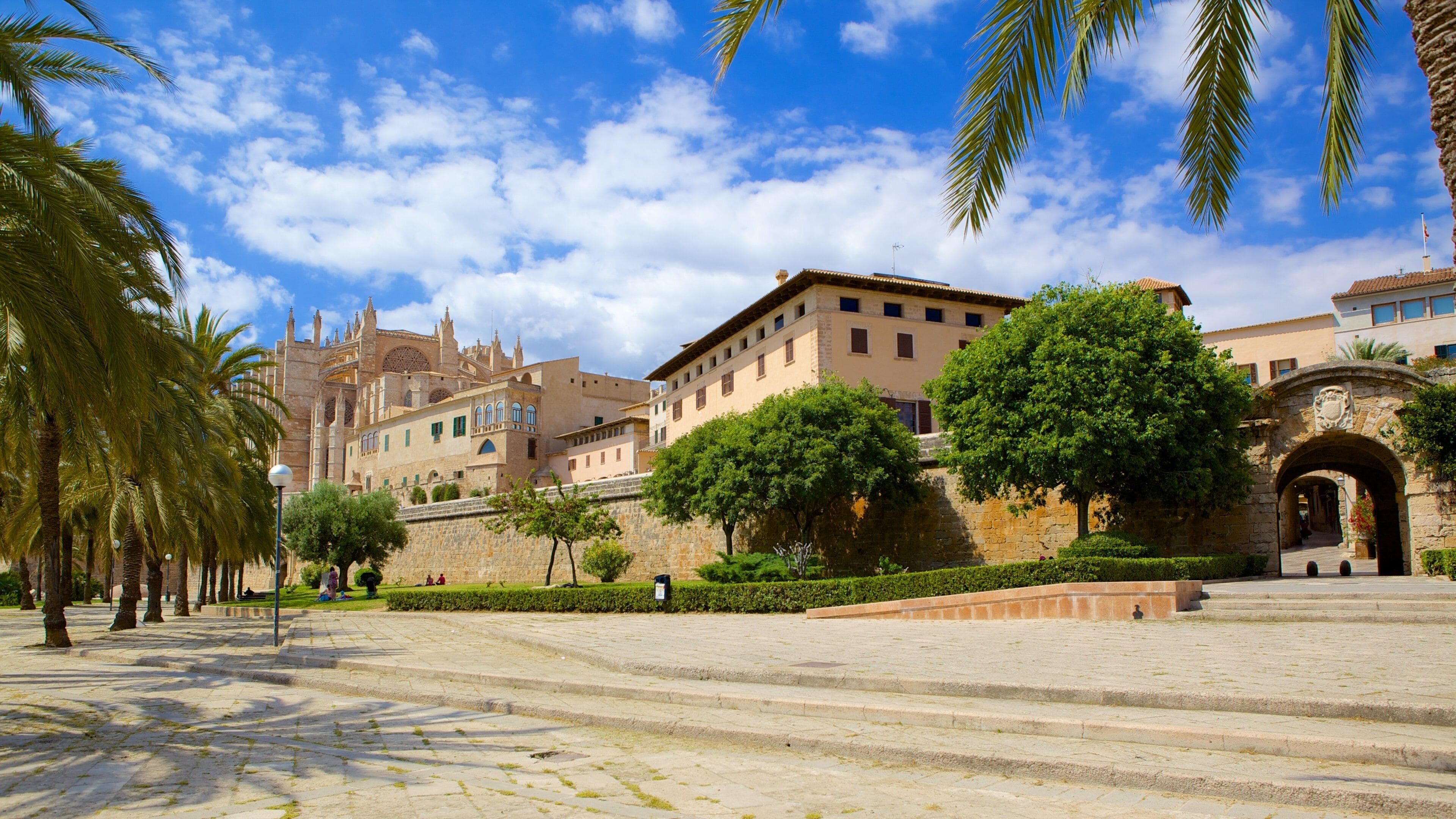 A historic square with cobblestone pavement, palm trees, and traditional buildings with arches and towers.