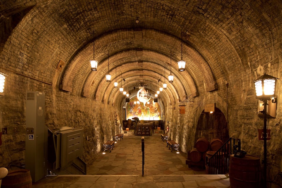 A stone-walled, arched corridor with hanging lanterns and wooden barrels.