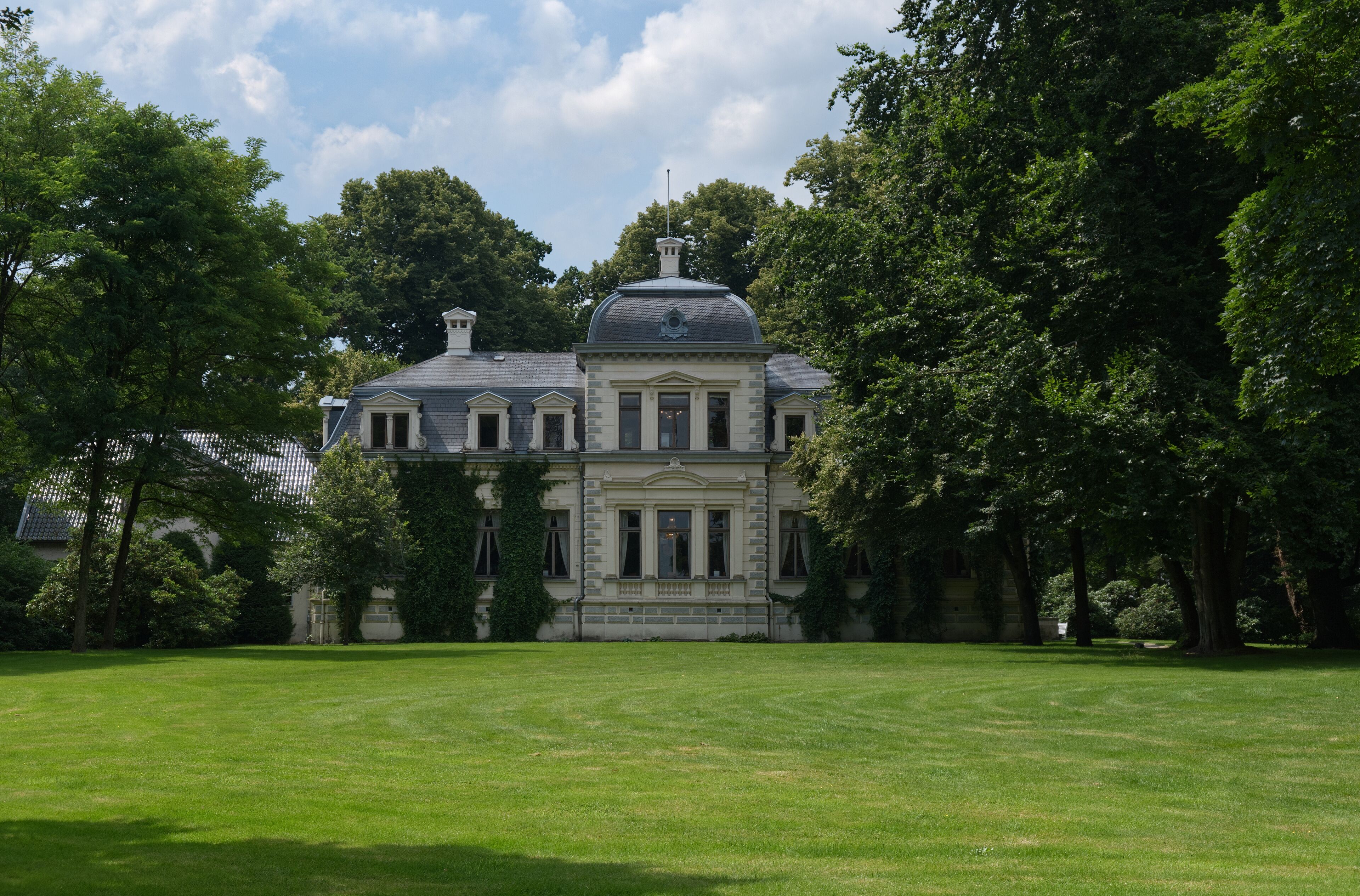A large, historic building with a prominent dome and columns, surrounded by lush green trees and a well-maintained lawn.