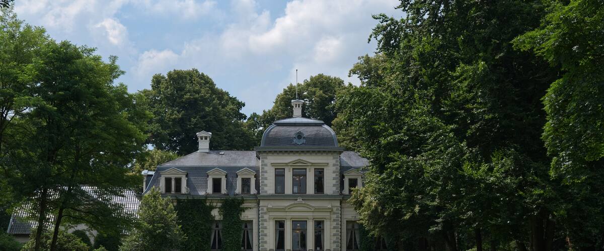A large, historic building with a prominent dome and columns, surrounded by lush green trees and a well-maintained lawn.