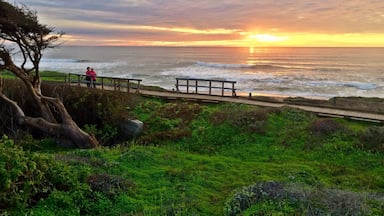 Um pôr do sol costeiro com uma passarela de madeira e pessoas observando o oceano.