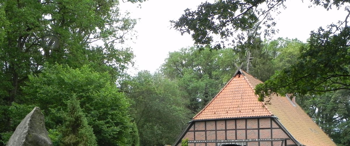 A traditional half-timbered house with a steeply pitched roof, surrounded by lush greenery and a large rock.