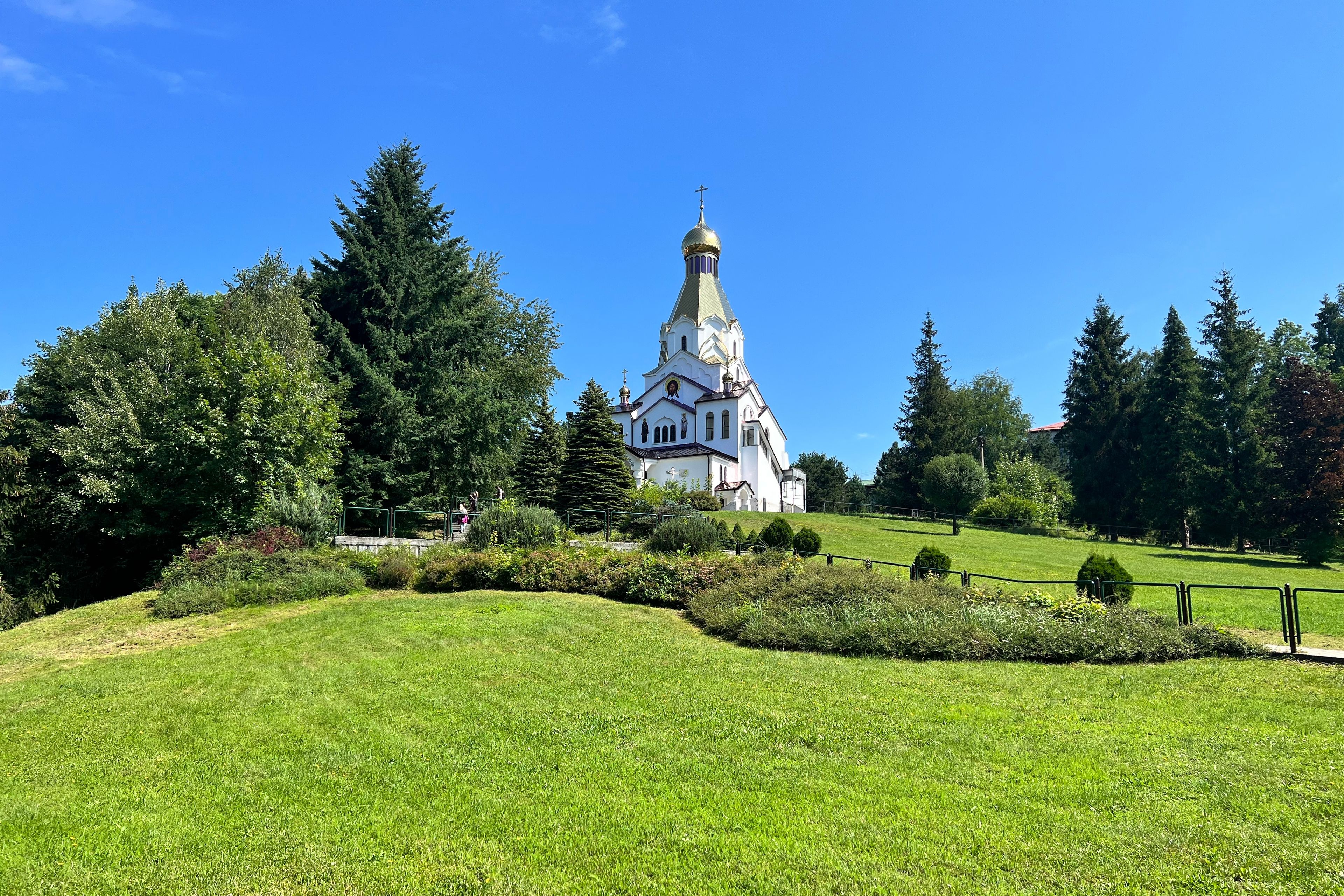 A white church with a tall spire and golden dome, surrounded by lush greenery and trees.