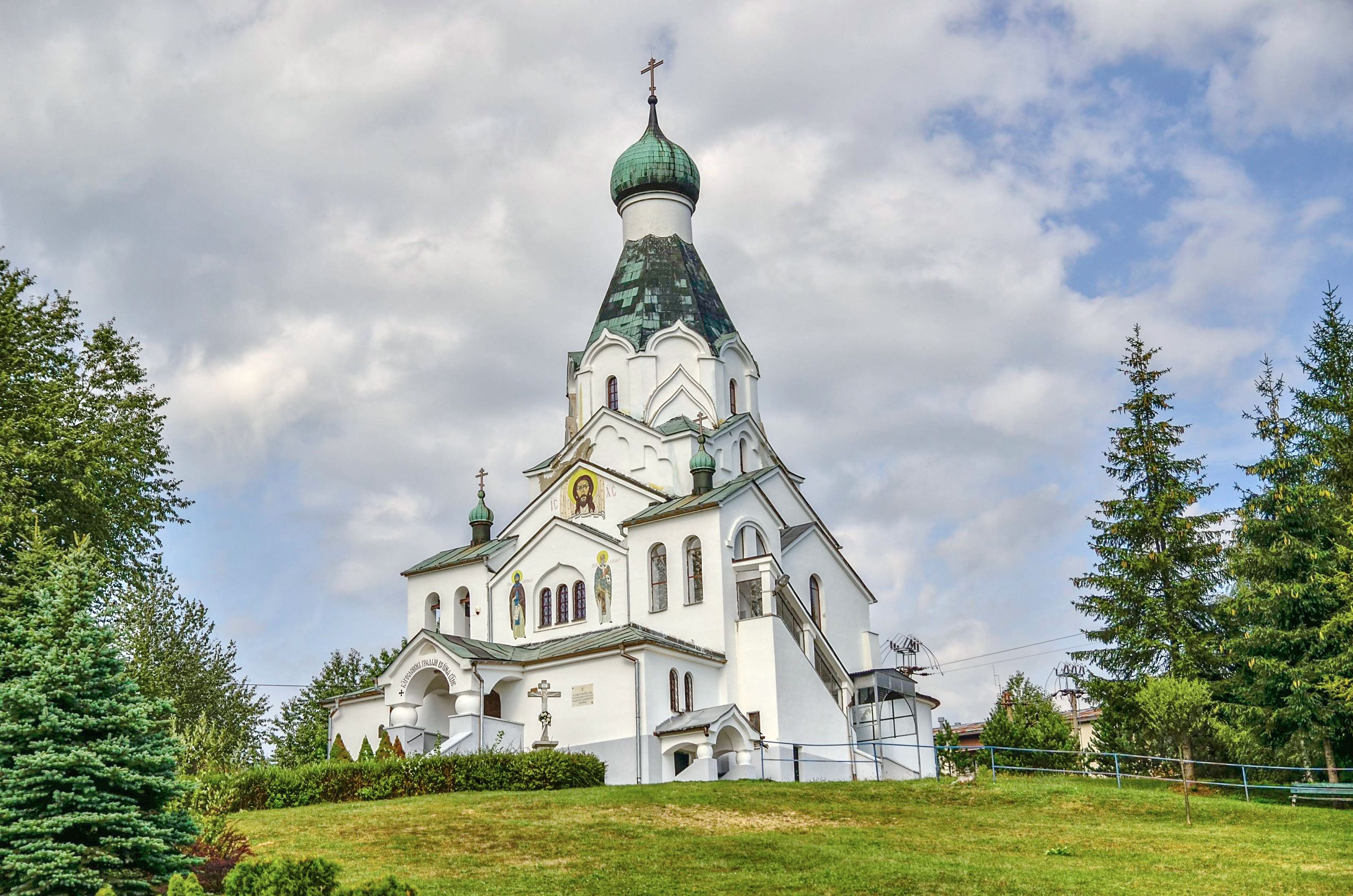 A white church with a green dome and crosses, surrounded by trees and grass.