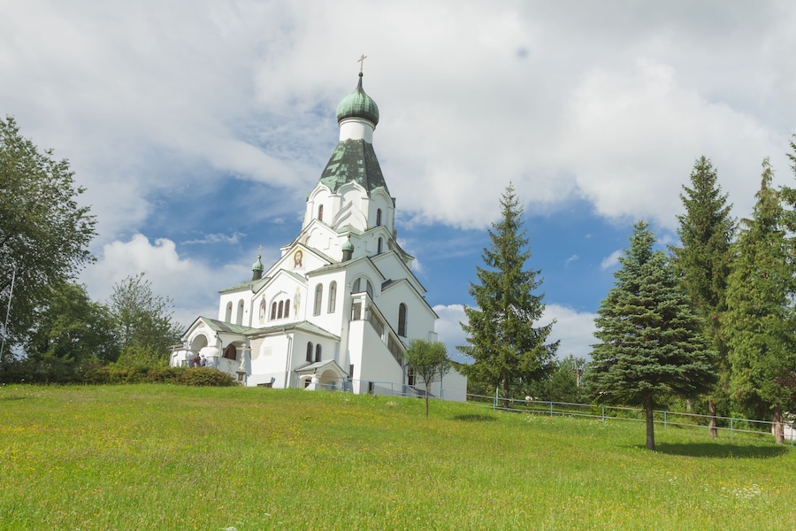 Uma igreja branca com cúpula verde e cruz, rodeada por árvores e grama.