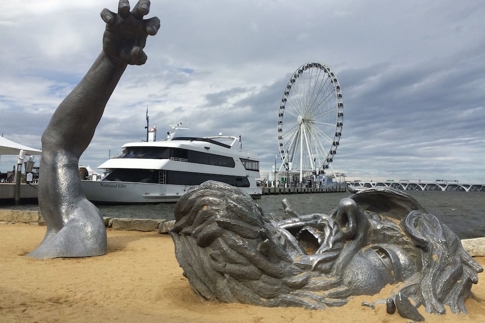 Une sculpture représentant une main tendue vers le ciel, une grande roue et un yacht baptisé « National Elite » sur une plage.