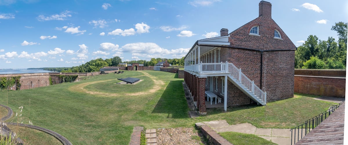 A brick house with a white porch and a staircase, surrounded by a grassy lawn and a brick wall.