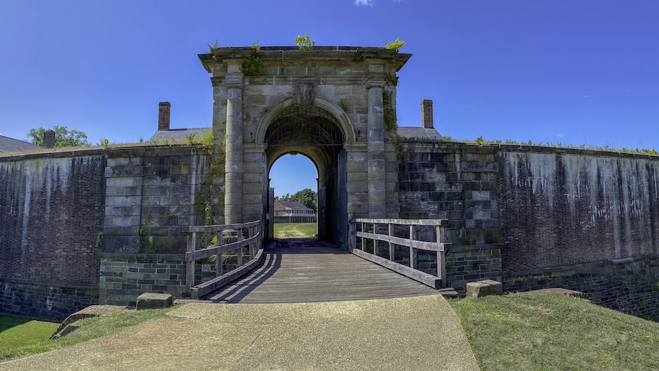 A stone archway leading into a historic fortification with a wooden bridge and a clear blue sky.