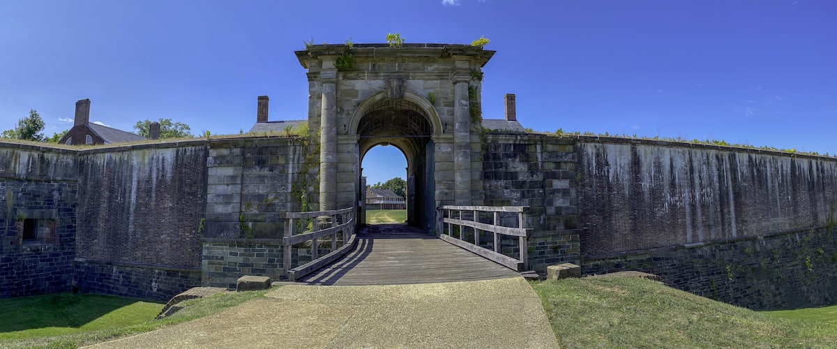 A stone archway leading into a historic fortification with a wooden bridge and a clear blue sky.