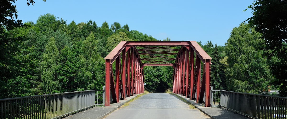 A red metal bridge with a metal railing, surrounded by green trees.