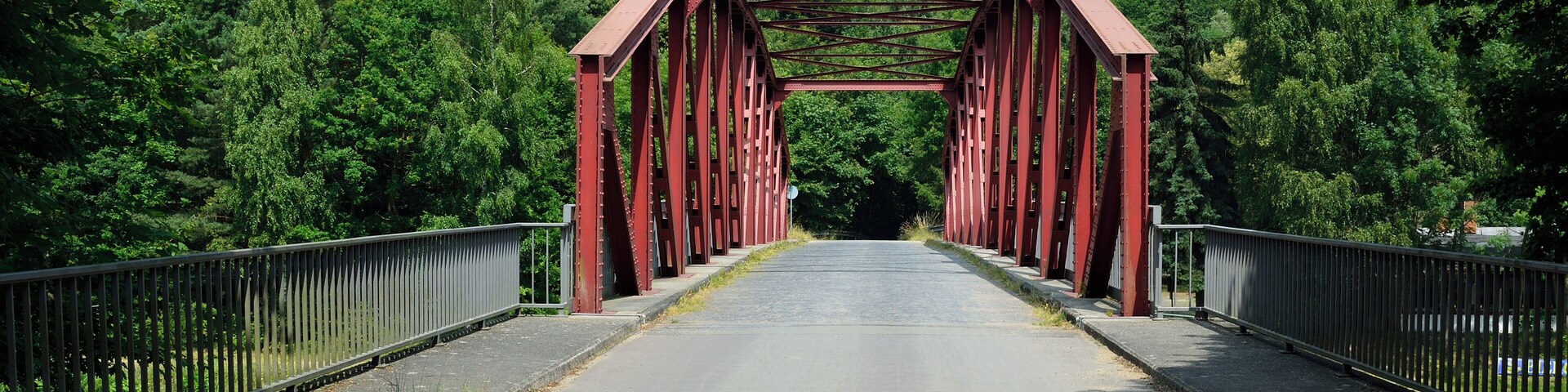 A red metal bridge with a metal railing, surrounded by green trees.