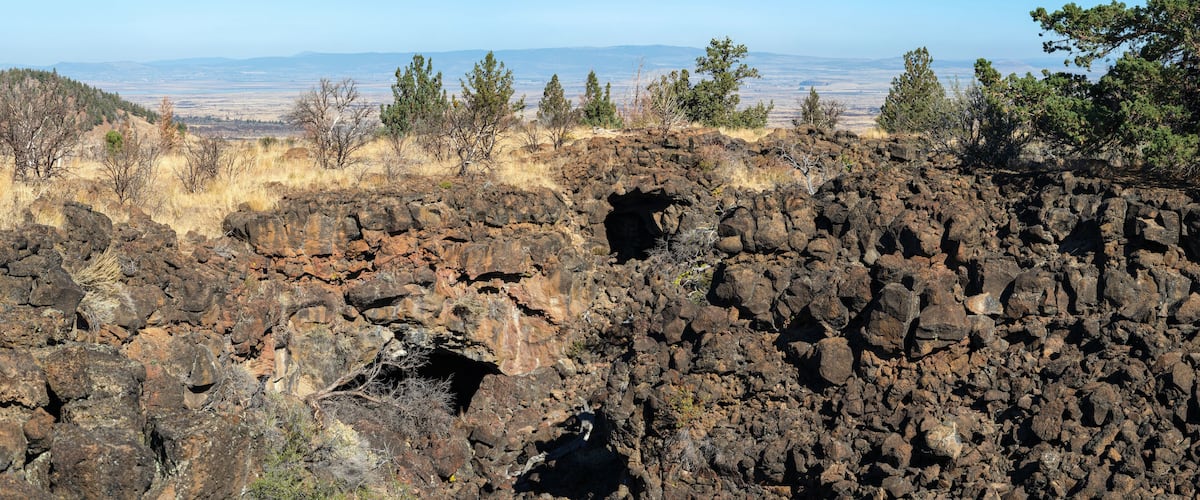 A rocky landscape with a large cave entrance, sparse vegetation, and distant mountains.