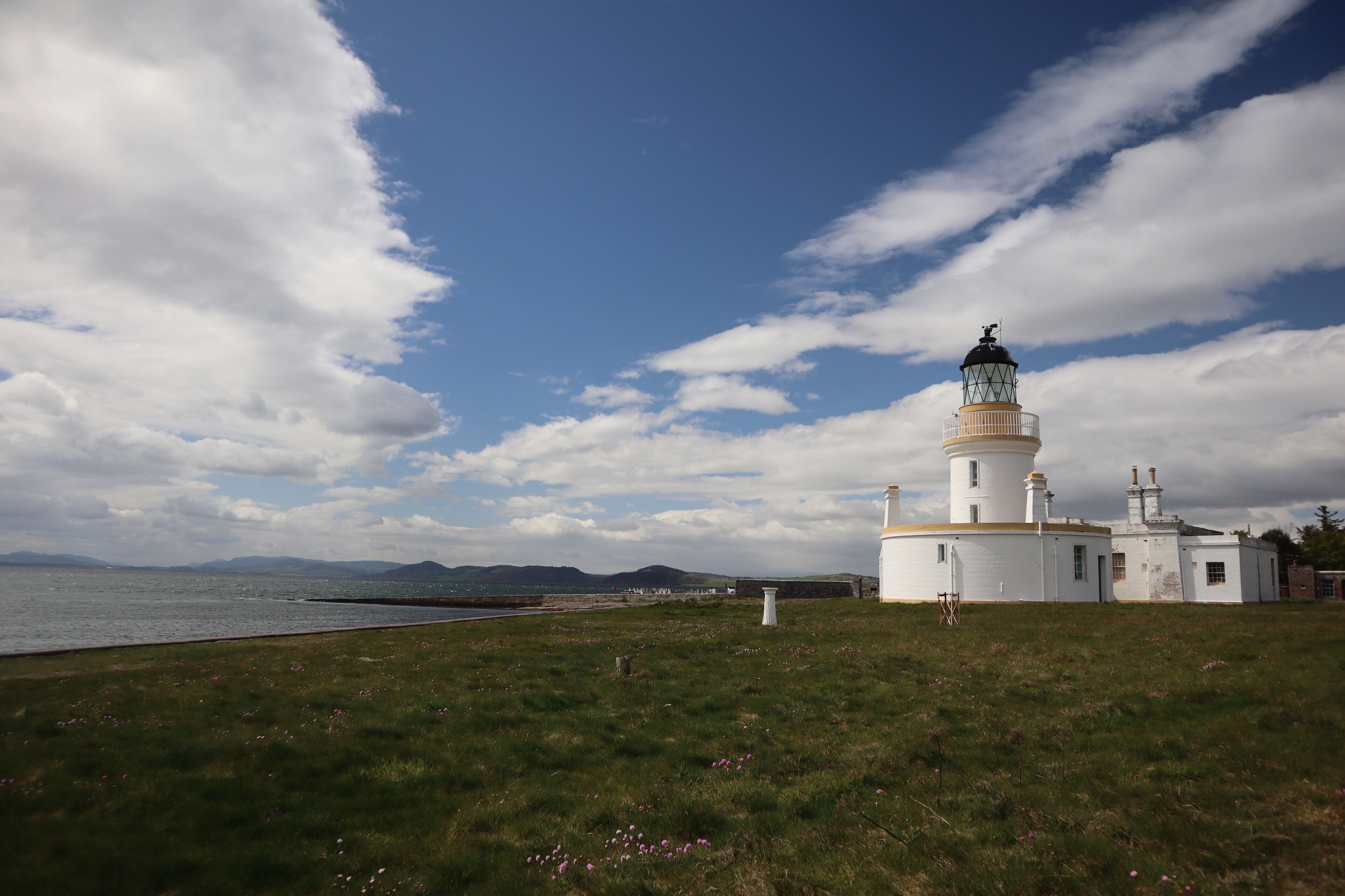 A lighthouse with a white exterior and a green top, situated on a grassy hill overlooking a body of water with mountains in the distance.