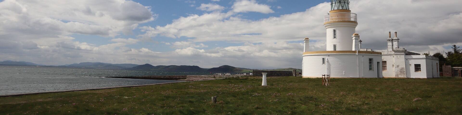 A lighthouse with a white exterior and a green top, situated on a grassy hill overlooking a body of water with mountains in the distance.